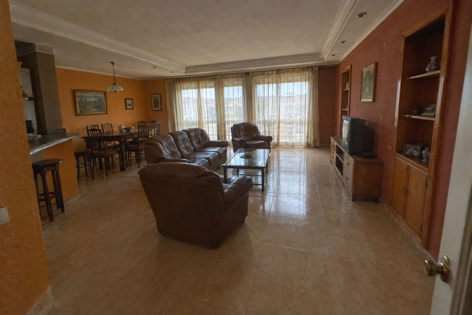 Salón amplio antes de pintar, con gotelé, paredes en ocre y rojo teja, muebles de piel y visillos